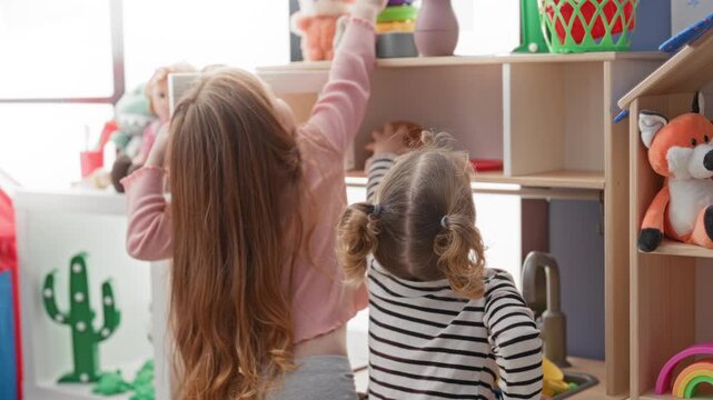 Two girls reach with hands for toys on a shelf in a bright playroom building while one grabs a stuffed fox and the other opens a toy cabinet; shared curiosity.