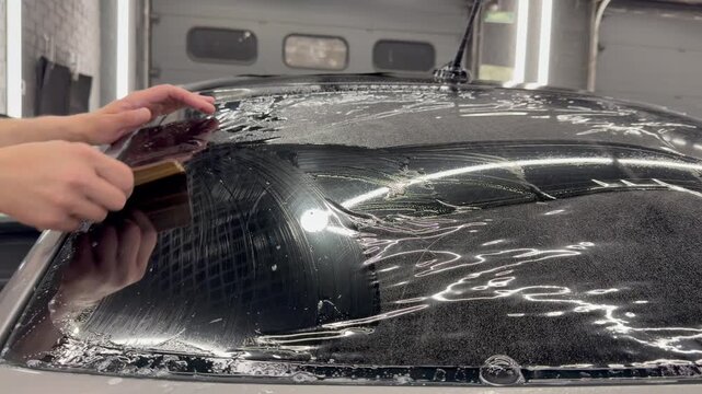 Professional car window tinting process: technician prepares the glass, applies tint film, uses a heat gun to shape it on curved surfaces, and smooths out air bubbles with a squeegee. 