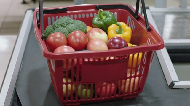 Man in white coat placing shopping basket with fresh vegetables and fruits on the checkout conveyor belt at the supermarket for payment processing at the register