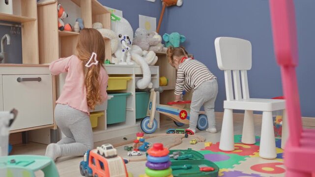 Two girls playing on colorful foam mats, one pushes a wooden scooter while the other holds stacking rings and a toy truck in a playroom inside a building; playful bonding.