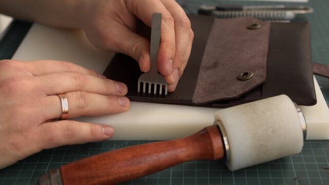 Craftsman punching stitching holes in brown genuine leather using a pricking iron and mallet for a handmade wallet