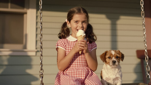 A happy young girl enjoys an ice cream cone with her cute dog on a sunny porch swing. This joyful childhood scene captures summer fun and friendship