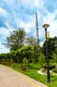 Parks and cell tower in a green space. People walk along a path in a park with trees and flowers under a cloudy sky. A cell tower stands in the background.