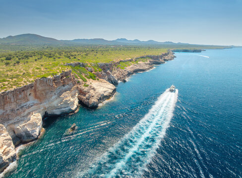 Aerial view of cruise boat moving near rocky cliffs of Mallorca, Spain. Passenger boat with white foam wake trail in blue Mediterranean sea, coastal rocks. Drone summer landscape. Top view. Yacht