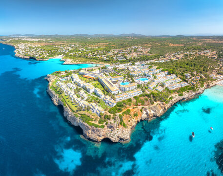 Aerial drone view of sea coast with white resort houses on rocky cliffs in summer. Turquoise blue sea water, sunny sky, boats, sandy beach. Luxury travel destination, Mallorca island, Spain. Top view