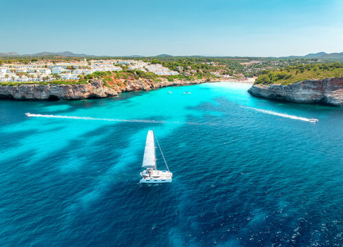 Aerial view of sailing catamaran yacht in turquoise sea water bay. Sailboat. Mallorca, Spain. Luxury boat cruising in ocean cove near sandy beach, resort houses on rocky coast. Drone summer seascape