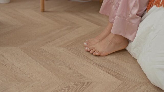 Woman on bed in pink pyjamas places bare feet on herringbone wooden floor, toes curling with heel touching the floor beside white duvet in bedroom; morning calm.