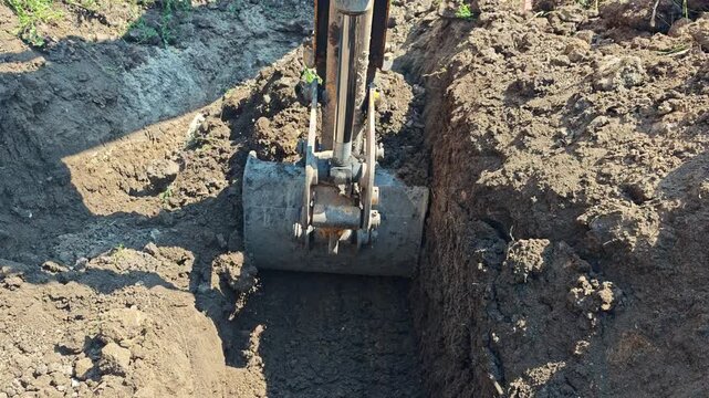 Yellow excavator bucket tamping and flattening loose soil at the bottom of a narrow trench.