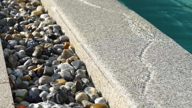 A close-up, selective focus shot of a luxury overflow swimming pool edge made of granite stone, with clear water running over it down into a drainage channel decorated with river pebbles.