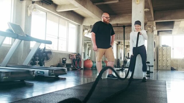 In a sunlit gym, a woman works battle ropes while a man watches, surrounded by treadmills, exercise balls, and lockers