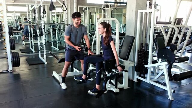 Two people in a gym training session; a woman uses a hip abductor machine while a trainer offers guidance among weight machines and mirrors.