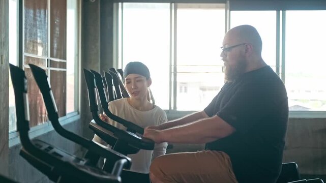 Trainer guides a man on a stationary bike in a sunlit gym, adjusting the console during a cardio workout.