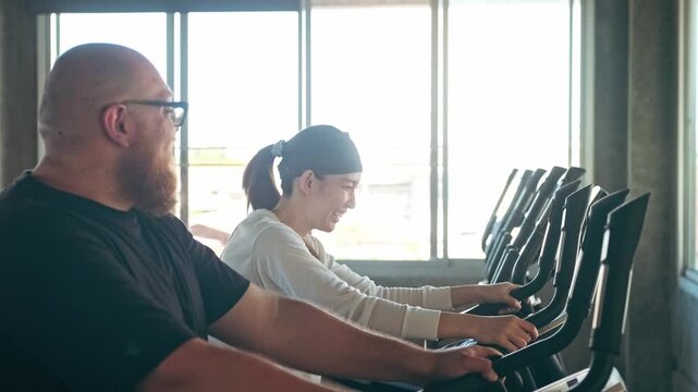 Two people chat and smile while pedaling on exercise bikes in a bright gym, enjoying a cardio workout beside large windows.