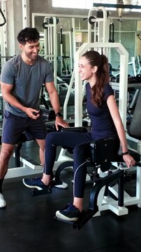 A woman uses a seated hip abductor machine while a fitness trainer supervises in a modern gym filled with weight equipment.