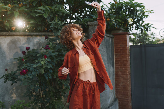 Young woman with curly hair dances outdoors in a rust jacket and matching pants, lifting an arm toward the sun in a casual, carefree moment