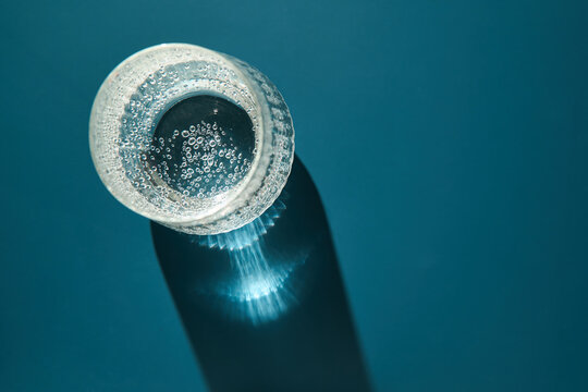 Glass Bottle Top With Effervescent Water And Visible Bubbles Casting Long Radial Light Shadow On Smooth Blue Surface In Studio Minimal Still Life Composition.
