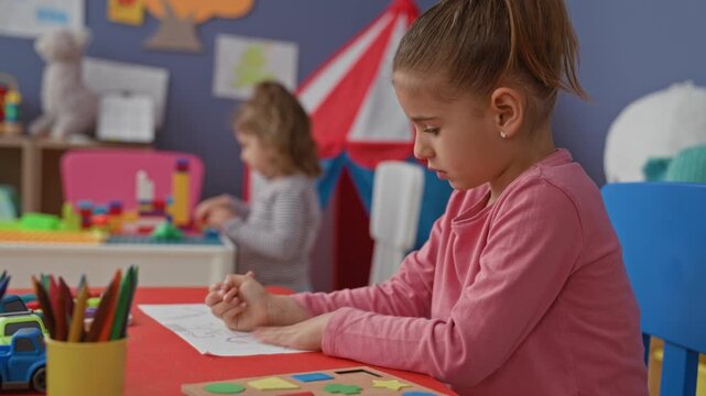 Girl reaches with hand for a colored pencil on a red table in a preschool classroom building, drawing and sorting crayons with toys and shelf behind her; quiet concentration.
