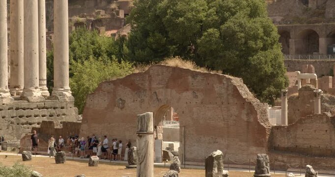 A group of tourists explores the iconic Roman Forum, walking past massive ancient columns and a large weathered brick wall structure, surrounded by green trees on a bright summer day in Rome, Italy.