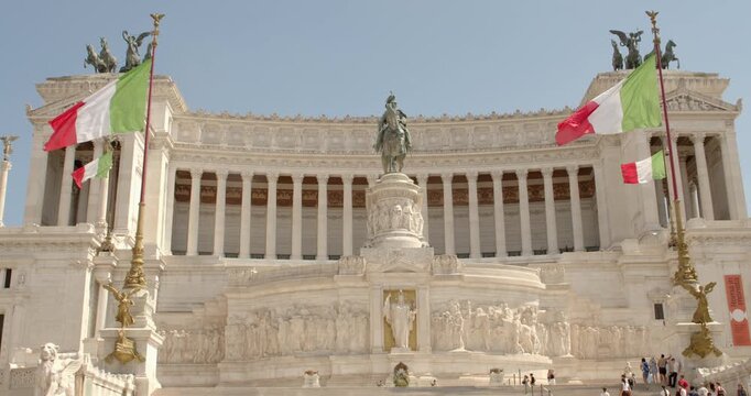 View of the majestic white marble Altare della Patria monument in Rome, Italy. The panorama is dominated by the central equestrian statue and numerous Italian flags waving under a bright summer sky.