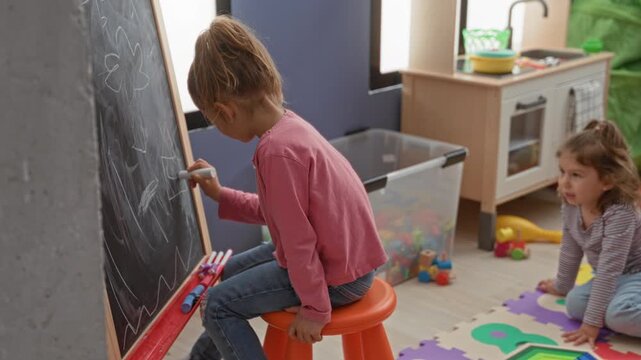 Girl writes on chalkboard with chalk and hand visible while another child watches on playmat inside a building; playful learning.