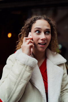 Surprised woman looking away with wide eyes during smartphone call, emotional reaction to unexpected mobile notification or news, candid photography of expressive female user in city