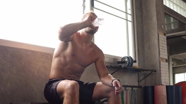 Muscular athletic man drink water from bottle inside gym. Sweaty male athlete take break during fitness workout training for muscle recovery
