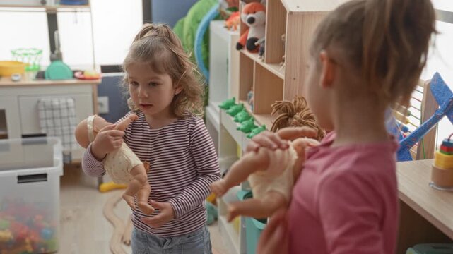 Two girls holding dolls, one pointing finger while the other cradles a baby doll in a preschool playroom; playful bonding.