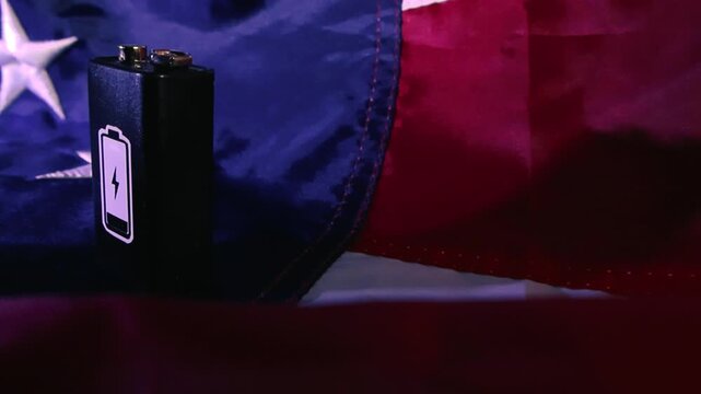 Macro shot of a battery with a charge symbol placed on a waving American flag, conceptually representing national energy policies, economic struggles, and the country's power resources