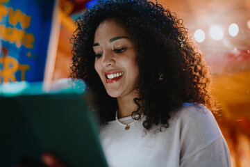Happy young woman with curly hair looking down at smart tablet device screen in cozy indoor cafe, creative freelancer working remotely or female student e-learning online concept portrait © BullRun