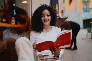Beautiful young woman holding open red book and looking at camera with happy smile on cozy cafe terrace, female student studying outdoor, youth education and lifestyle concept portrait © BullRun
