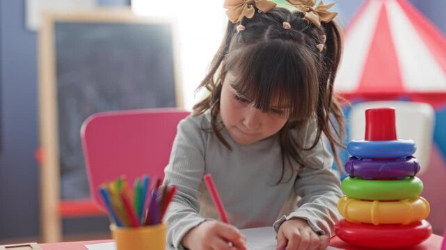 Girl drawing with pink pencil, right hand holding pencil while coloring on paper at preschool classroom playroom table with markers cup and stacking rings nearby; concentration creativity.