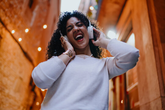 Joyful young woman touching wireless headphones and laughing out loud while walking on cozy evening street, happy female tourist enjoying music or audio guide, youth lifestyle portrait