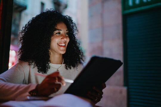 Smiling young woman holding digital tablet and stylus pen in outdoor evening cafe, creative freelance designer enjoying online remote work process and learning