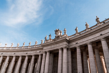 Naklejka premium Curved classical colonnade with stone statues atop tall columns against a bright blue sky with wispy clouds