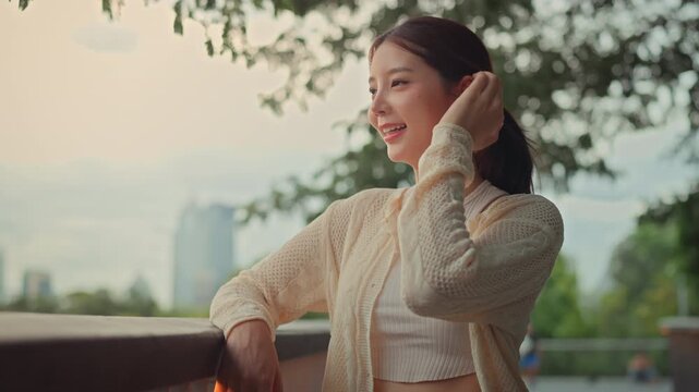 Happy Asian woman smiling and tucking hair behind ear while relaxing outdoors, beautiful young female traveler looking away at green city park with blur skyscraper background for urban lifestyle