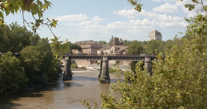 View of the muddy Tiber River in Rome, Italy, showing the historic Palatino Bridge framed by lush green trees, with traditional buildings and the prominent synagogue dome visible under a sunny sky.