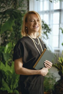 A confident woman stands in a vibrant indoor garden, smiling while holding a green book. Her casual yet elegant outfit and pearl necklace complement the lively plants around her