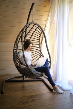 Young woman sitting by the window in a hanging swing chair with a laptop on her knees
