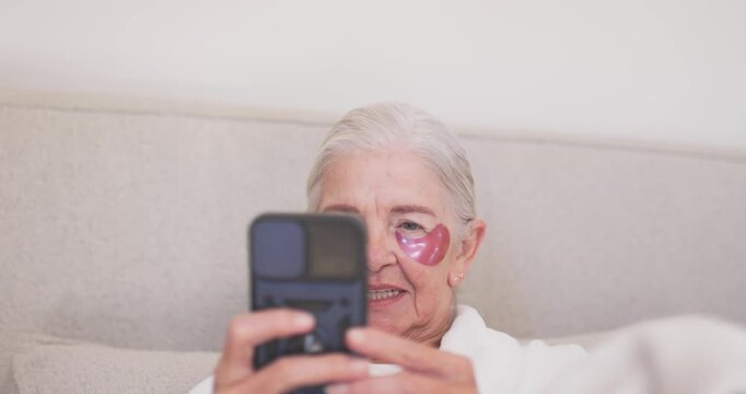 Senior woman on couch wearing robe pink heart patches checking phone adjusting camera for selfie