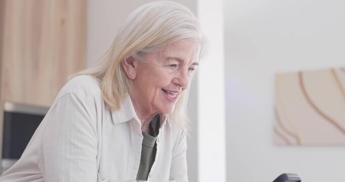 Senior woman glancing at smartphone, tapping and scrolling messages in kitchen with glass