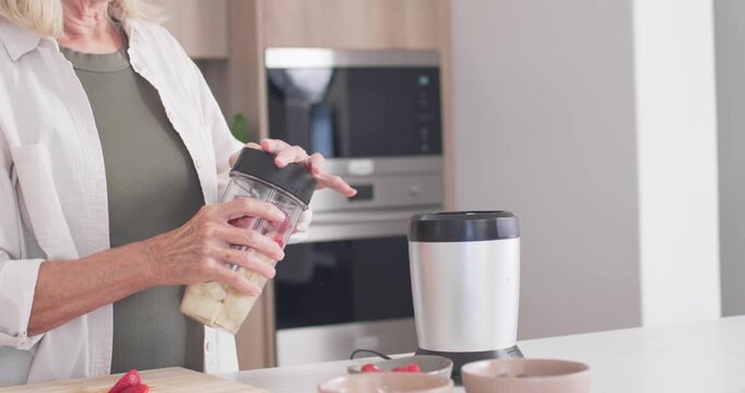Senior woman holding blender cup with fruit securing lid inverting cup onto blender base at counter