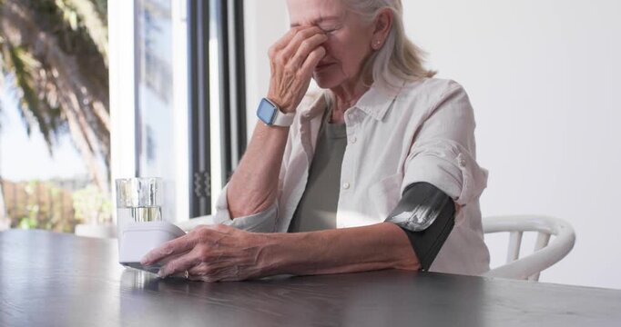 Senior woman sitting at table by glass door, resting on monitor, rubbing nose, checking arm cuff