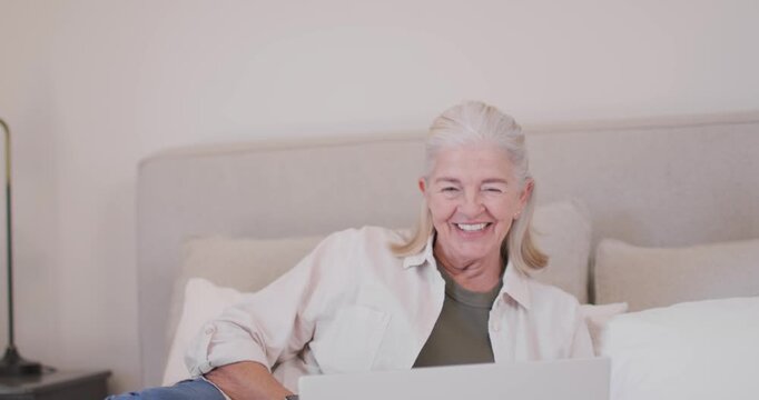 Senior woman reacting to laptop content, laughing then smiling, speaking and adjusting pose on bed