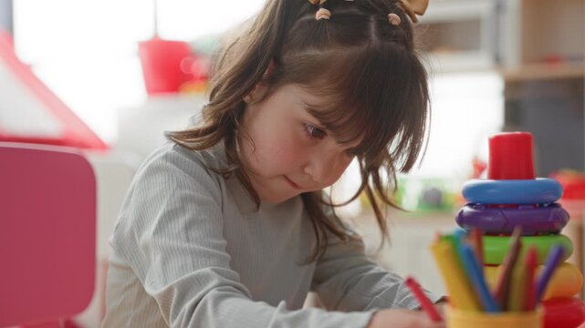 Girl drawing with colored pencils beside a stack of plastic rings on a low table inside a bright playroom building; creative concentration.