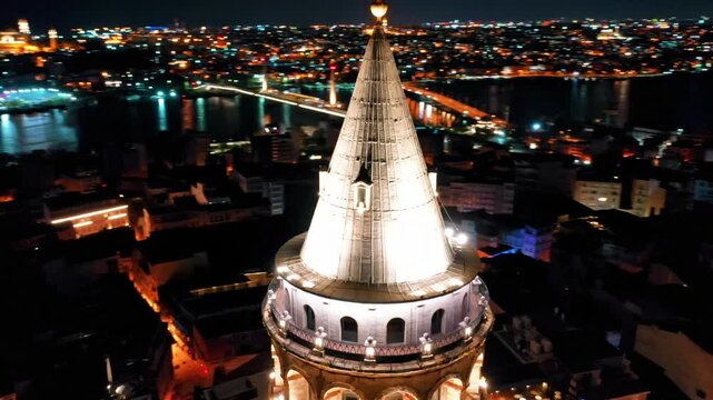 Galata Tower aerial drone shot in Istanbul at night. Panoramic view of illuminated historic landmark, Bosphorus and city lights in darkness.