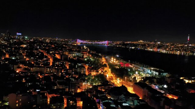 Galata Tower aerial drone shot in Istanbul at night. Panoramic view of illuminated historic landmark, Bosphorus and city lights in darkness.