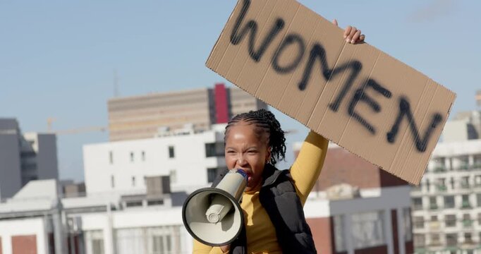 African American woman in yellow top raising WOMEN sign, speaking into megaphone for women on roof