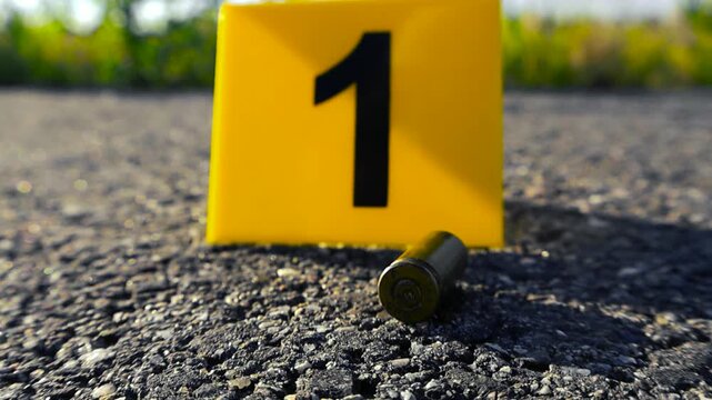 Yellow forensic evidence marker with the number one and a spent bullet casing laying on the asphalt at a crime scene, showcasing a police investigation into a violent shooting incident