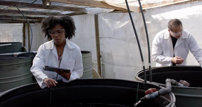 Researchers sampling tanks in shed, woman dipping probe recording on tablet, senior man lifting net