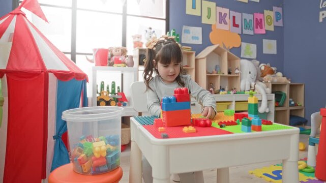 Girl stacking plastic toy blocks with hand on a play table in building with shelves and tent; curiosity concentration.